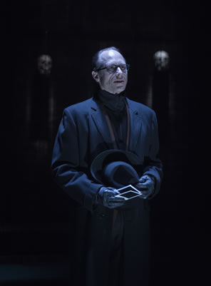 Tyrrell in black overcoat, black turtleneck, wearing glasses and  wearing gloves as he holds his black fedora hat in his left hand and two poloroid prints in his right.