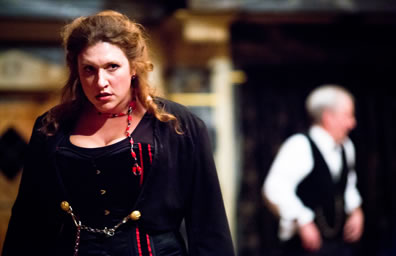 Kate in Victorian black dress with red streaks and red and blue braids scowls while in blurry background stands Petruchio in black vest and white shirt
