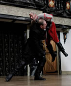 Richard all in black lifts Somerset, in gray shirt and red knee pagts, into the air on the tip of his sword.
