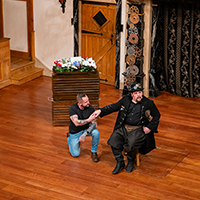 A man in black t-shirt and jeans and lots of tattoos on his arm kneels on one knee holding the extended hand of Malvolio in black steampunk outfit sitting on a stool in the middle of the stage. Behind them is two stacked crates topped by a tray of flowers.