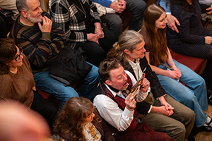 Justin McCombs as Andrew Aguechek sits with a playbill up to his face squeezed in between two audience members on a crowded cushioed bench.