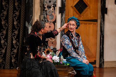 Angelique Archer as Olivia in a long black dress with black jacket embroidered with flowers and Summer England as Viola disguised as Cesario wearing a coloruflly flowered shirt, purple vest, royal blue pants and blu cap sit on each side of a trunk with flowers between them. Olivia is reaching across to stroke Cesario's temple as Viola/Cesario looks uncomfortably to the audience.