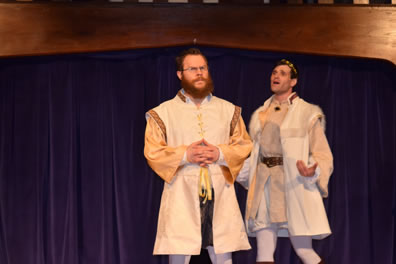 Leontes to the right with hands cupped and outstretched in exasperation talking to the side of Camillo, who patiently holds his hands folded at his waist, both where white sleeveless tunics with satiny shirts underneath and white leggings, Leontes in a crown.