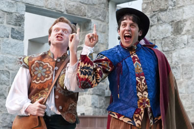 Tranio in brown flowery vest and blond hair, Lucentia in floral patterend hacket with blue vest, a hat and cape, both with fingers pointed in the air and "ah-Ha!" expressions