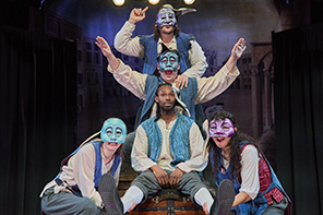 The 2024 Capulet cast in blue vests, all in masks except Romeo, form the Queen Mab visual: Mercutio on top speaking, then Benvolio with arms outstretched,  Romeo listening,  two servants at the bottom.
