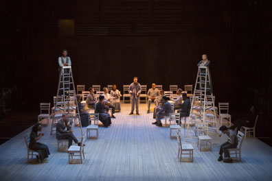 A platform with chairs, two ladders topped by George and Emily, their siblings seated in chairs below, his father, too, and toward the back of the stage other cast members sit around the choir director standing.