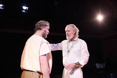 Timon in white nobleman's gown, gold medallion and wristbands talks gently with Flavius in simple shirt and pants and a headband