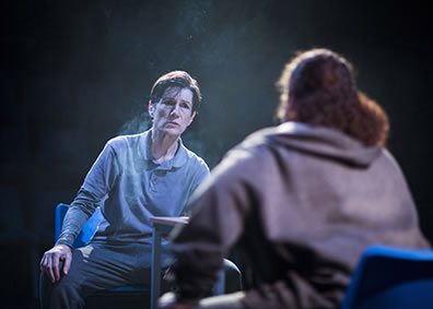 Production photo of Brutus sitting in prison clothes at a table talking with another prisoner