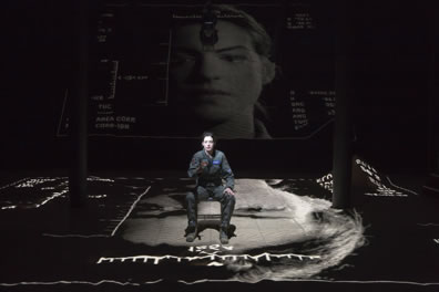 Hathaway in a flight suit sits on a chair in the middle of a gray-toned landscape with various coordinates on the edges, and a larger portrait of her projected on a screen in the background.