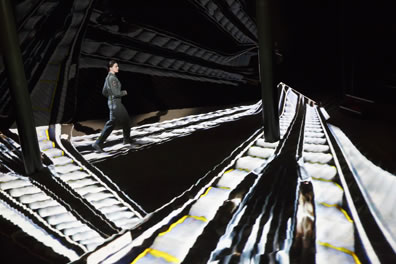 Hathaway, wearing a flightsuit, walking on an escalator, one of three criss-crossing the floor as projected on the sand