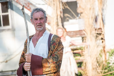 Prospero, wearing a cloak of symbol tapestry with large burgundy sleeves over a white shirt, he has a long stick clasped in both hands; behind him, Miranda is at the bottom of a reeds-lined fire escape against the white wall of an old building.