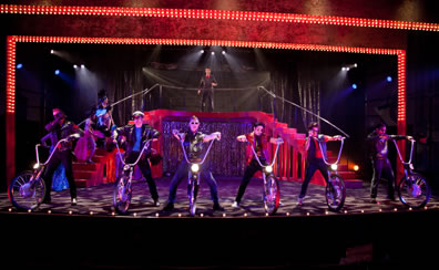 Lined up at the front of the stage, the biker stand with straddled legs, hands on the handlebars rising up from a single wheel and sporting a headlamp; a red double staircase leads to a platform where Leonato stands behind the rail; two showgirls stand on the left stair case. The silver gleamer curtain crosses the middle of the staircase, silver theater curtains are in the backdrop, and the stage floor is a pattern of black and white from the lighting.
