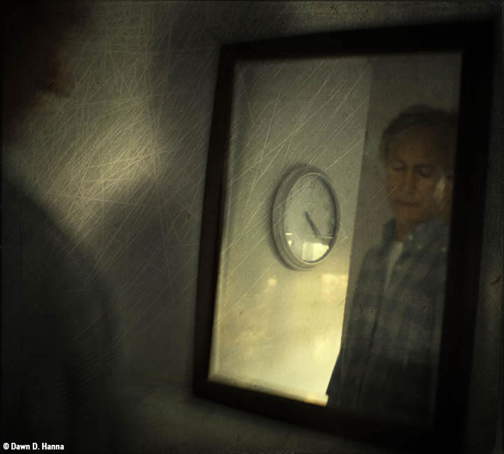 Reflection of a man in checkered work shirt in a mirror, he's looking down, and a clock on the wall is beside him in the reflection: time is 4:20; the shadow of his shoulder is to the left of the image, and the whole has a scratchy veneer